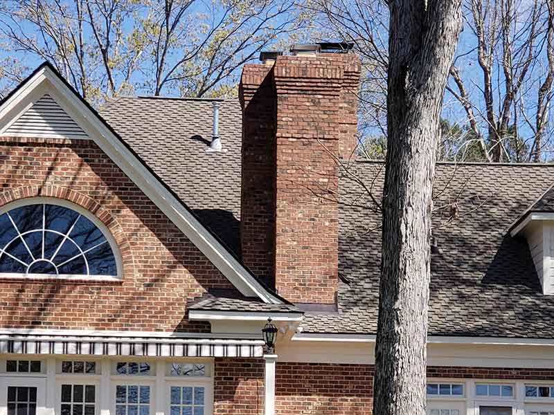 three tall masonry chimney exteriors with tree in the foreground and background a half round window in the peak of the home and lots of windows on the front of home