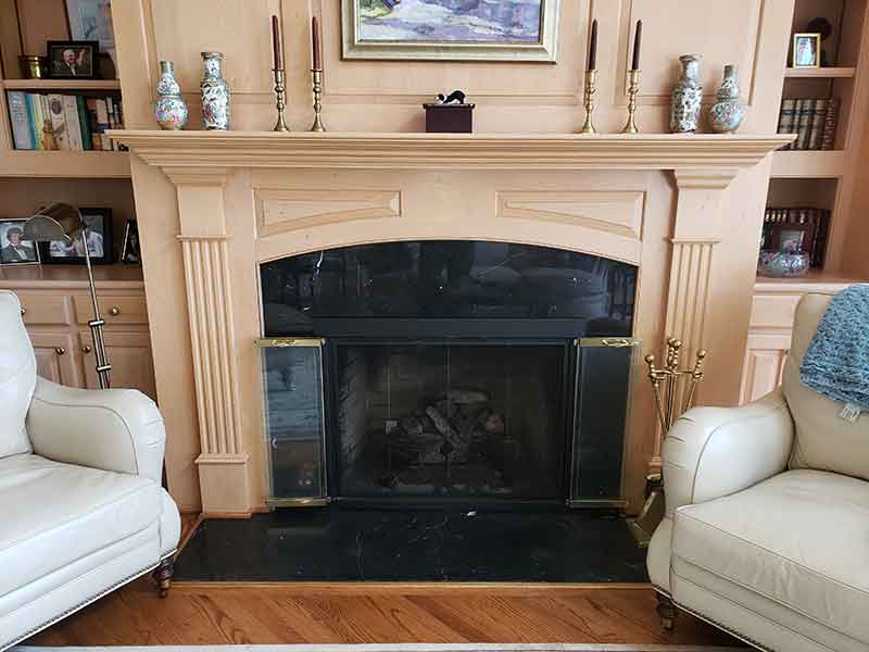 Beautiful beige color fireplace surround and mantel with folding glass doors mantel is decorated with candles and vases. Bookshelves and cabinets on each side with two white leather chairs on each side.