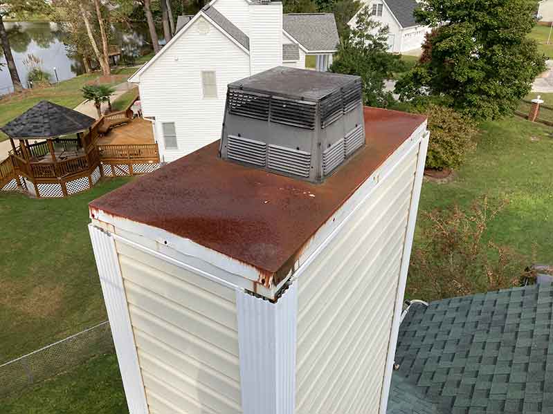 Vinyl sided chimney with rusted chase cover and metal chimney cap before chase cover replacement with homes in the background