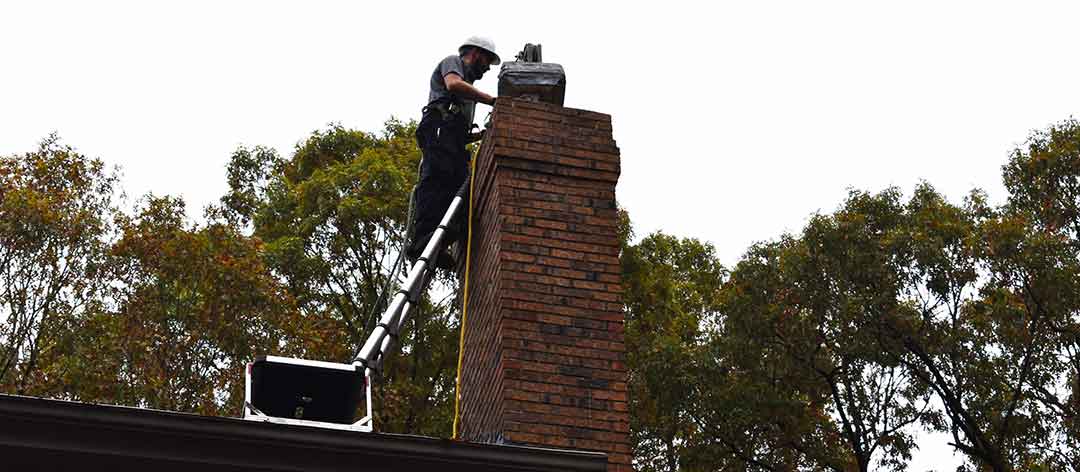 Chimney technician using specialized chimney inspection equipment while standing on a ladder to reach top of chimney on roof