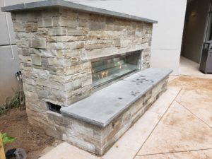 Left side view of completed custom outdoor fireplace made with natural cut stone, decorative glass and stone seating area/hearth