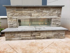 Front view of completed custom outdoor fireplace made with natural cut stone, decorative glass and stone seating area/hearth