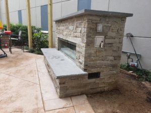 Right side view of completed custom outdoor fireplace made with natural cut stone, decorative glass and stone seating area/hearth