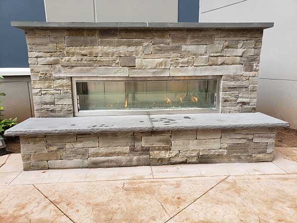 Mr Smokestack Original Photo of Custom Outdoor Fireplace - Front Front view of completed custom outdoor fireplace made with natural cut stone, decorative glass and stone seating area/hearth